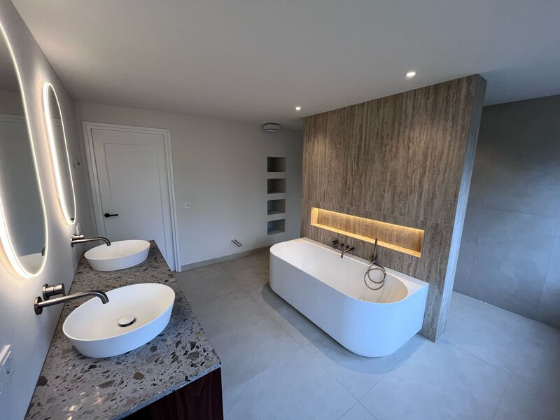 Ruime badkamer met vrijstaand bad, travertijnen wand met verlichte nis en dubbele wastafel in de verbouwde woning aan de Oranjelaan in Hilversum